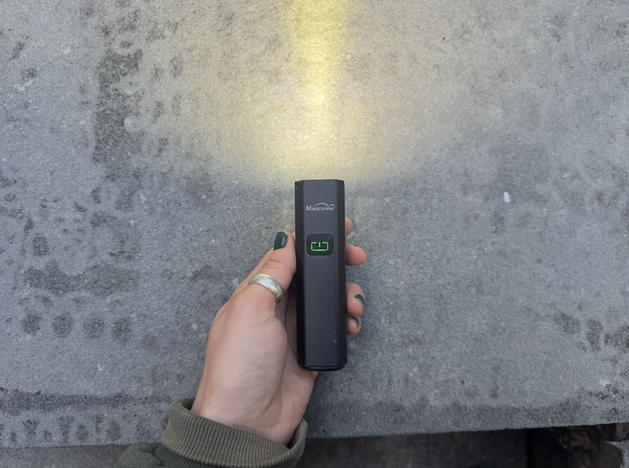 On a grey background, a hand holds a small square torch style bike light with a green illuminated on button and a yellow light. 